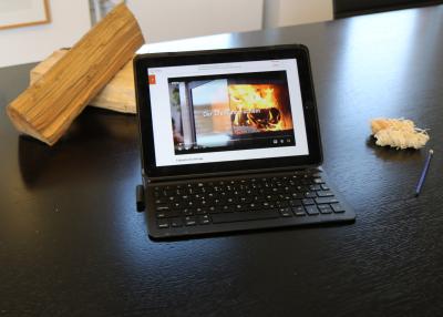 Schwarze Tischplatte darauf mittig ein mobiles Endgerät (Tablet) mit Tastatur davor, auf dem Bildschirm wird ein Video angezeigt zum "Ofenführerschein" mit Feuer in Kamin, rechts neben Gerät zwei Kaminanzünder und ein Stift, links hinter dem Gerät zwei helle Holzscheite übereinander