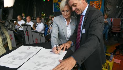 Bürgermeisterin von Beesel links unterschreibt neben Bürgermeister Frank Gellen eine von zwei Urkunden am Stehtisch auf Bühne vor Musikern